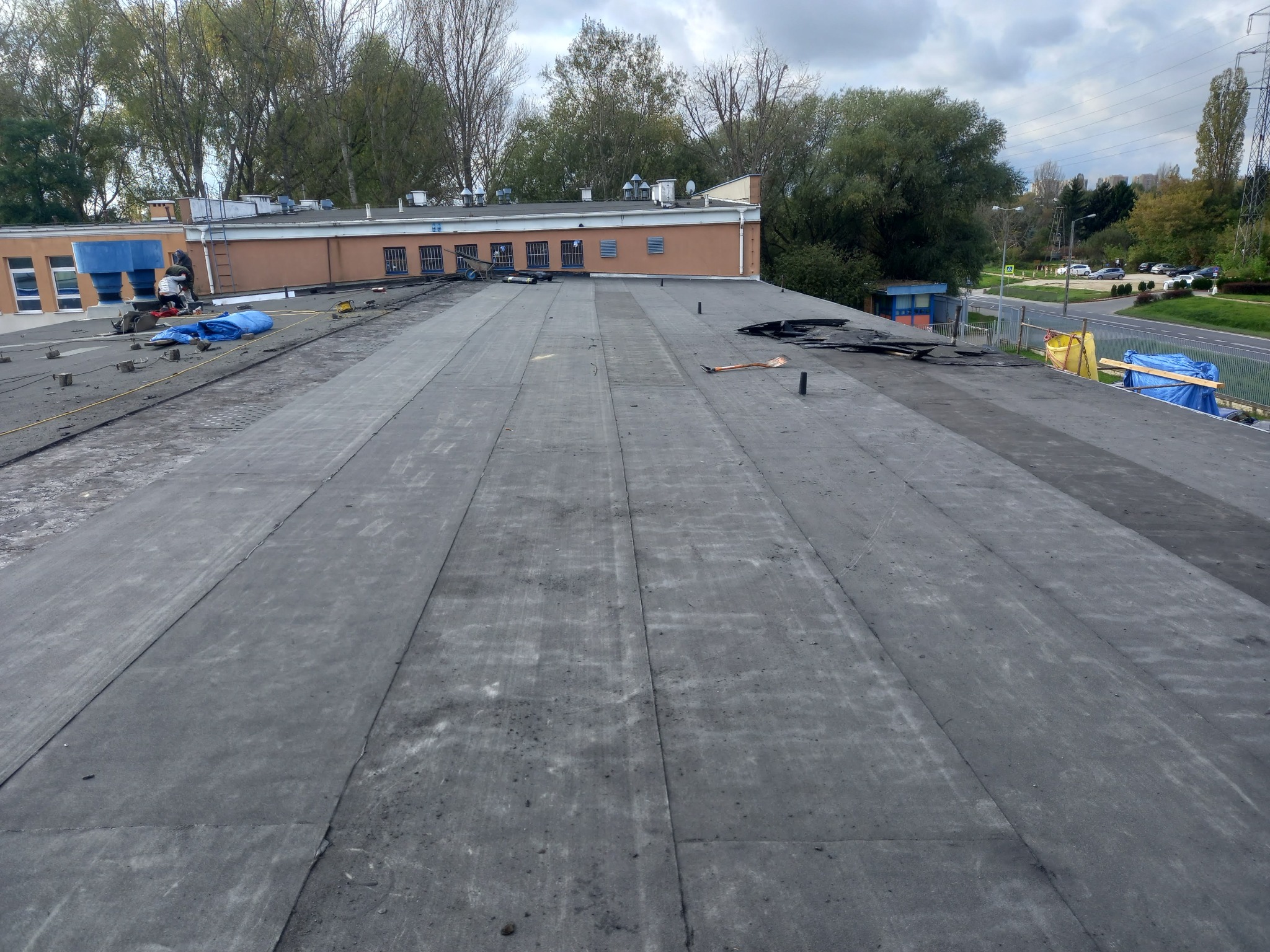 Widok z góry na płaski dach pokryty papą, z ekipą dekarzy pracującą w tle przy krawędzi budynku, narzędzia i materiały budowlane rozłożone na powierzchni dachu. W tle widoczne drzewa i budynki.