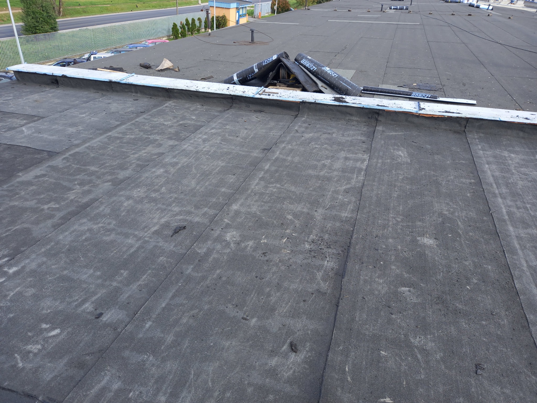 Widok z góry na płaski dach pokryty papą, z widocznymi rolkami papy IZOBUD i narzędziami przygotowanymi do pracy.