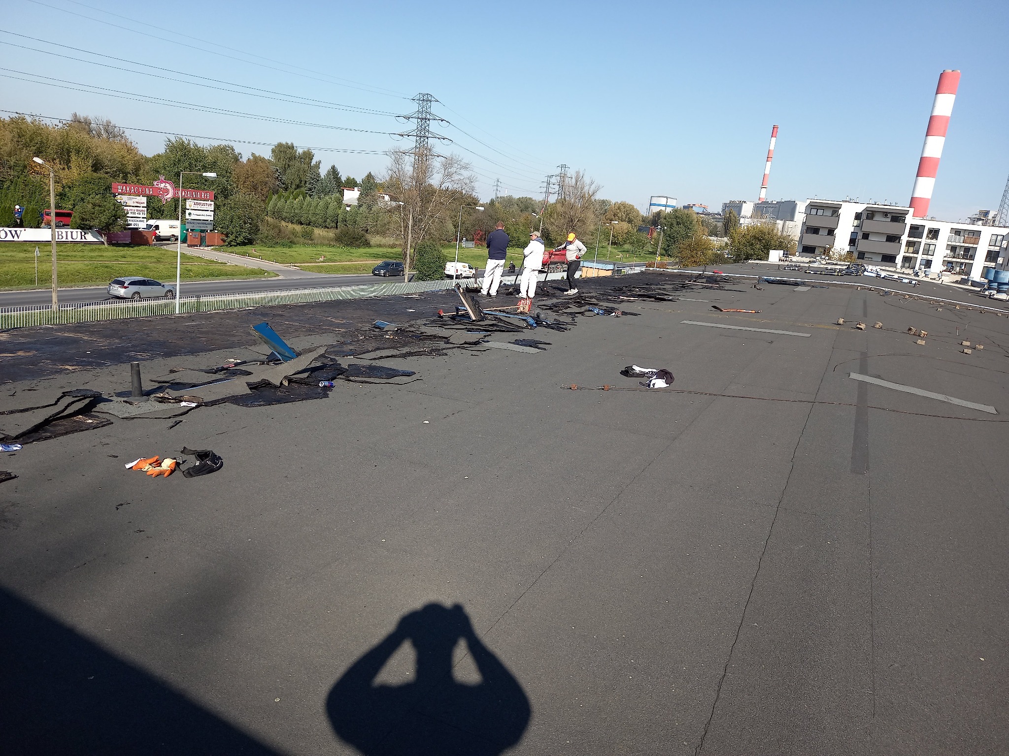 Ekipa dekarzy w trakcie demontażu starego pokrycia bitumicznego na dachu płaskim budynku przemysłowego. W tle widoczne kominy przemysłowe i budynki mieszkalne.