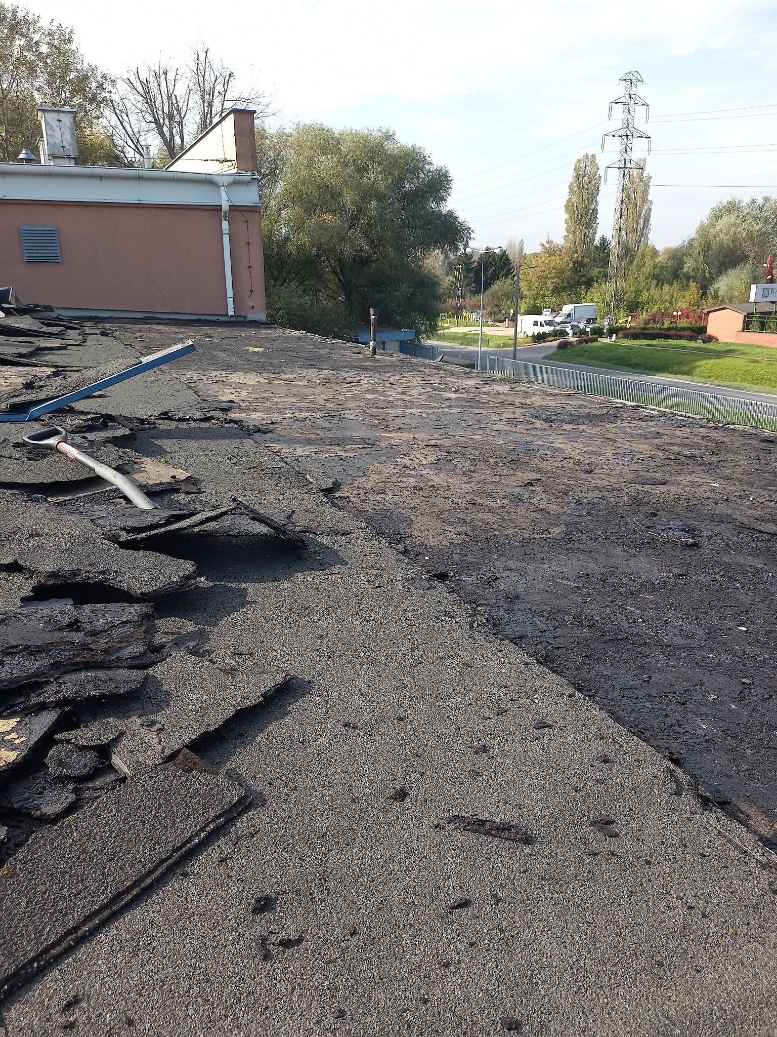 Demontaż starego pokrycia dachowego z papy, widoczne fragmenty odspojonej izolacji, przygotowanie pod nowe krycie, w tle budynek i słupy wysokiego napięcia.