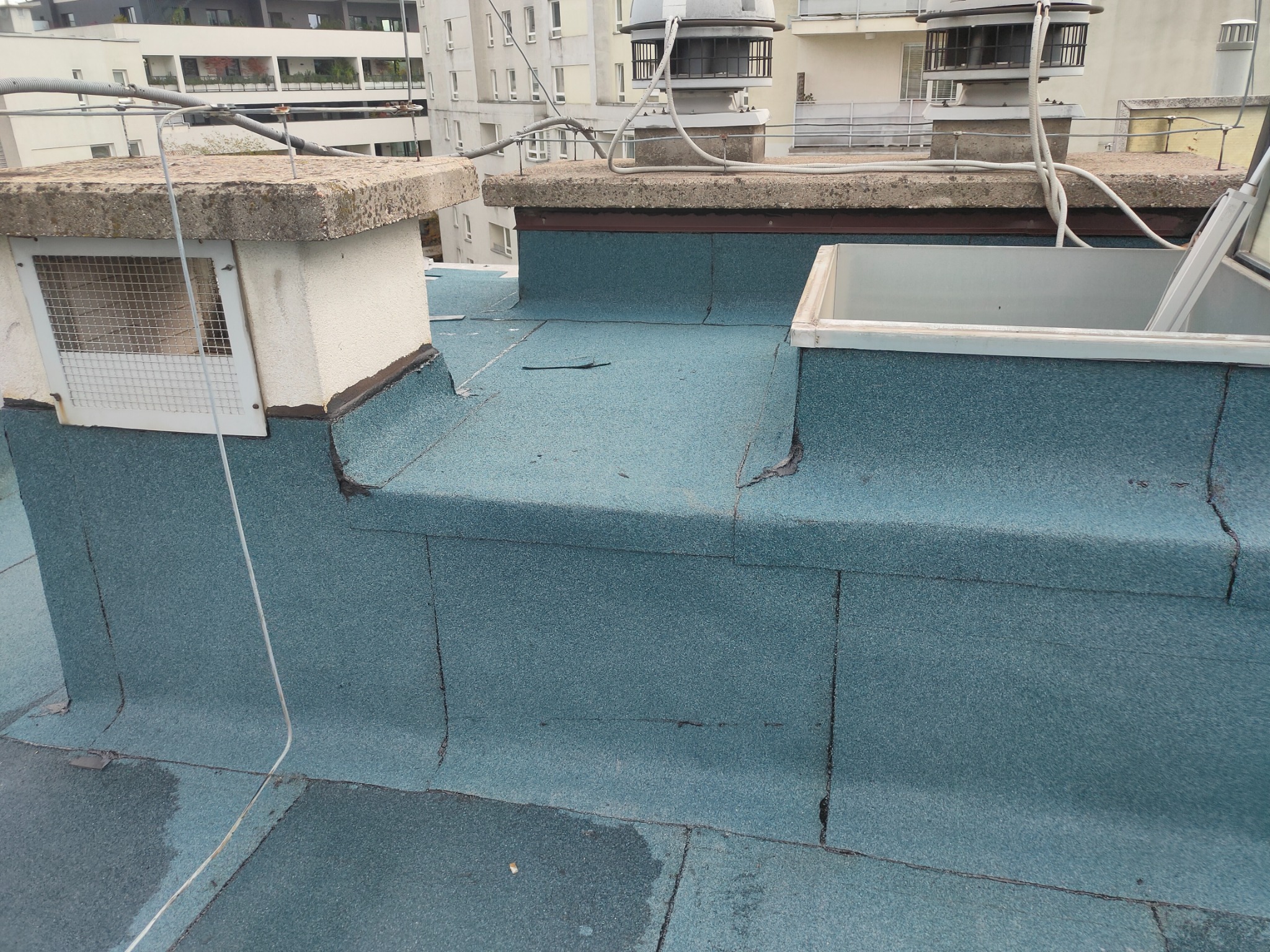 Niebieska papa na płaskim dachu z kominem wentylacyjnym i świetlikiem dachowym na tle zabudowy miejskiej.