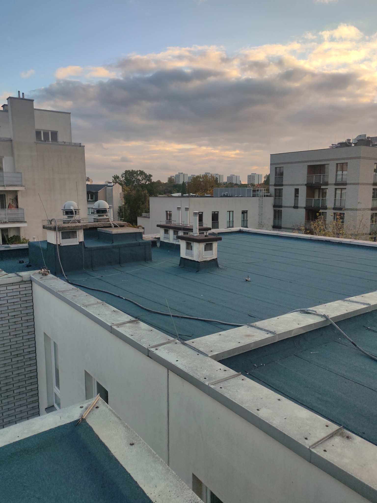 Niebieski dach płaski budynku wielorodzinnego z widocznymi kominami wentylacyjnymi i elementami obróbek blacharskich na tle zabudowy miejskiej i pochmurnego nieba.