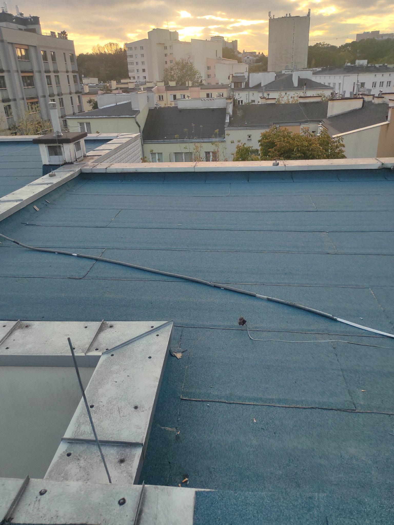 Widok z góry na płaski dach pokryty papą termozgrzewalną, z widocznymi elementami obróbek blacharskich wokół świetlika i przewodami na powierzchni, na tle zabudowy miejskiej i zachodzącego słońca.