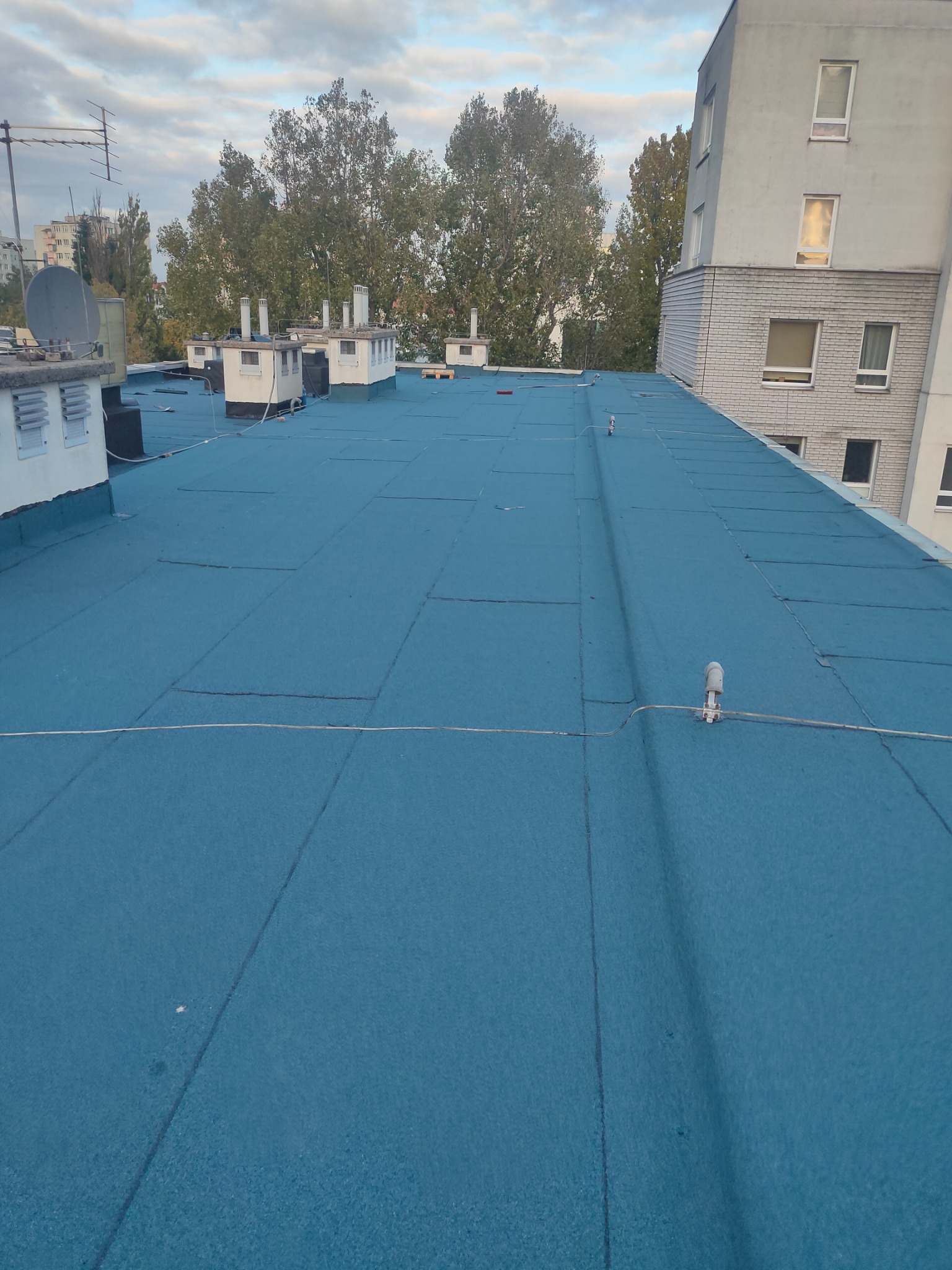 Niebieski, płaski dach pokryty papą termozgrzewalną na budynku wielorodzinnym, widoczne kominy wentylacyjne, anteny i elementy instalacji odgromowej.