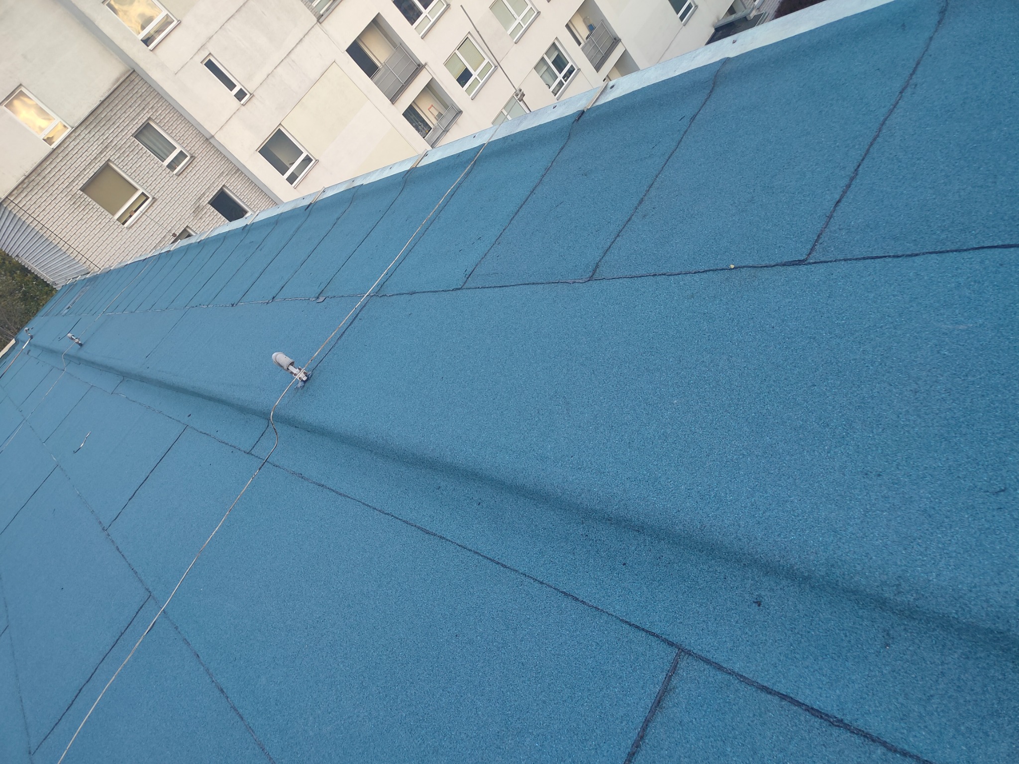 Niebieski dach pokryty papą termozgrzewalną na tle bloku mieszkalnego, widoczne przewody odgromowe.