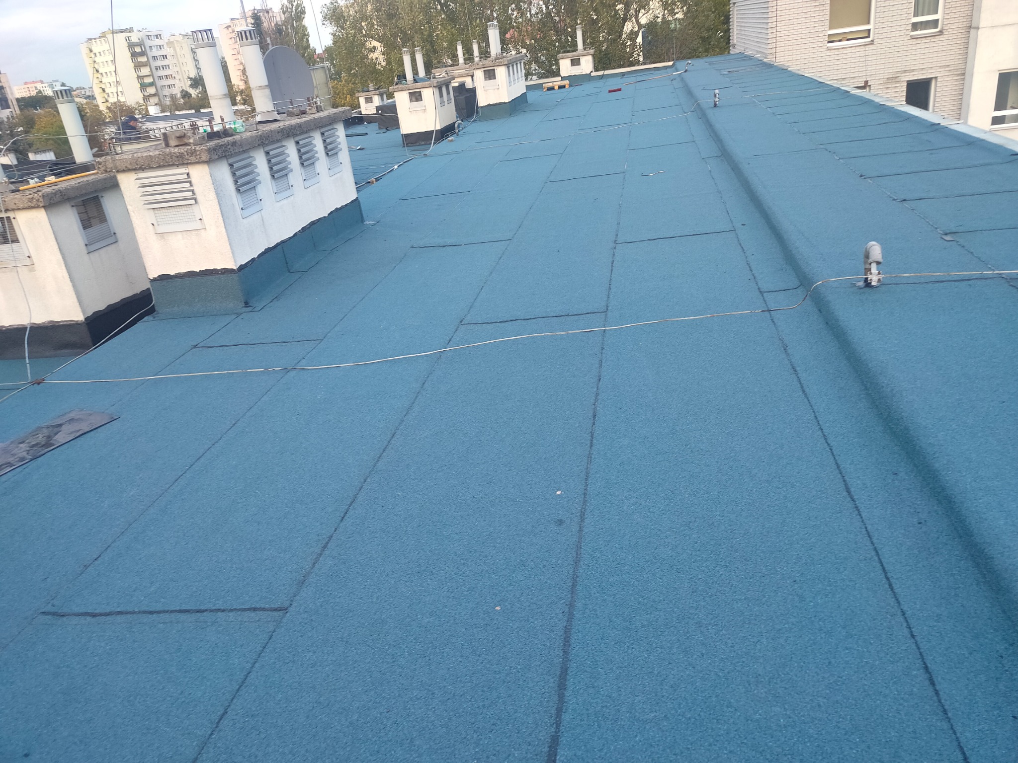 Niebieska, płaska powierzchnia dachu pokryta papą termozgrzewalną, widoczne kominy wentylacyjne i elementy instalacji odgromowej, w tle zabudowa miejska.