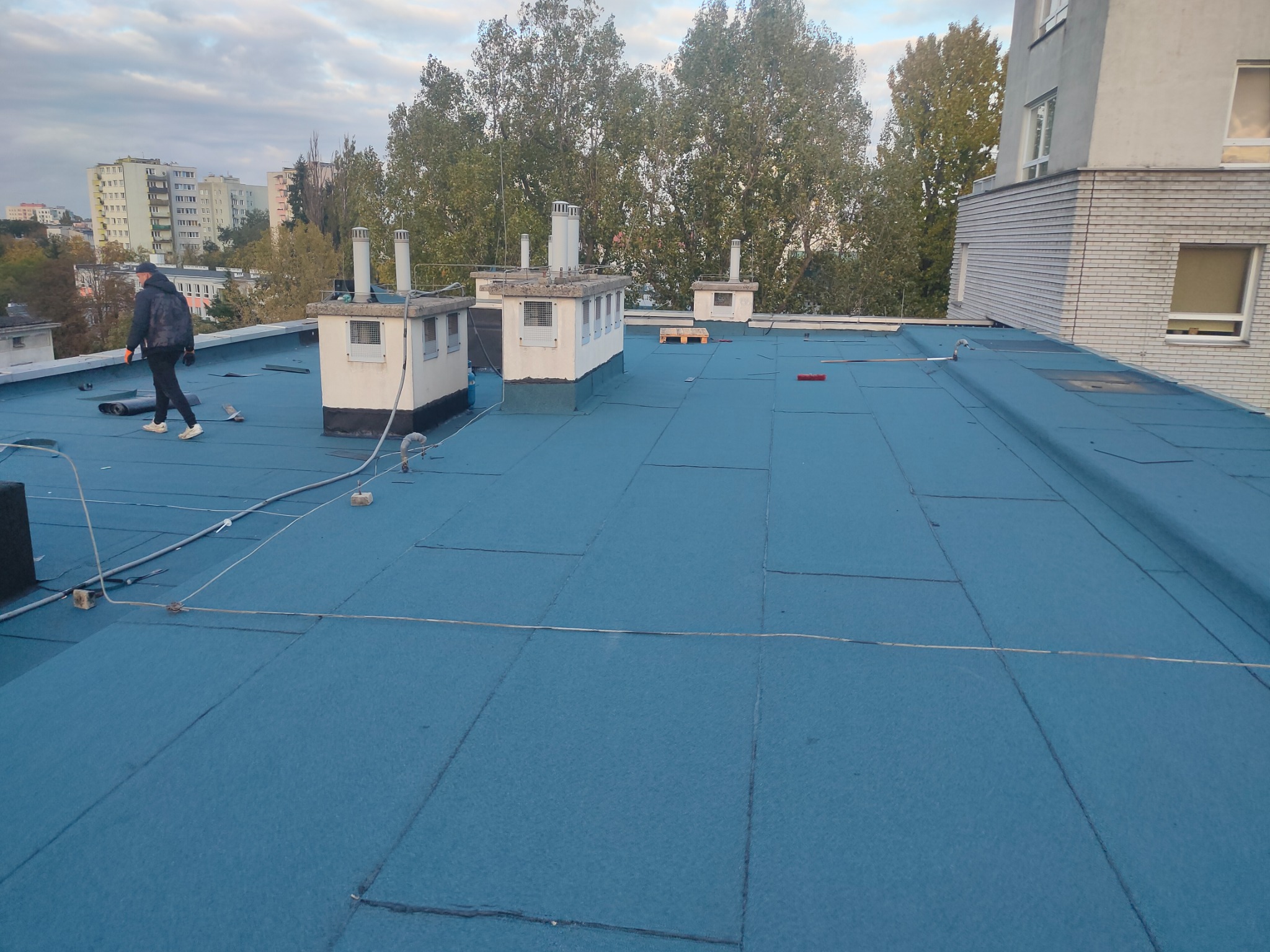 Niebieska płaska powierzchnia dachu pokryta papą termozgrzewalną, z widocznymi kominami wentylacyjnymi i sylwetką pracownika w oddali, na tle bloków mieszkalnych i drzew.