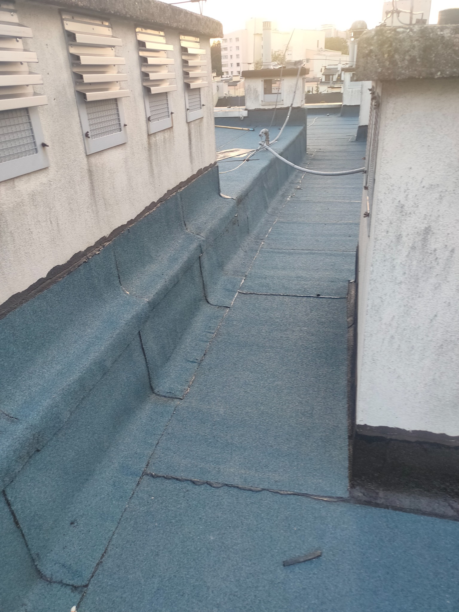 Płaski dach pokryty papą termozgrzewalną w kolorze niebiesko-szarym, widoczne łączenia papy i fragmenty ścian z otworami wentylacyjnymi.