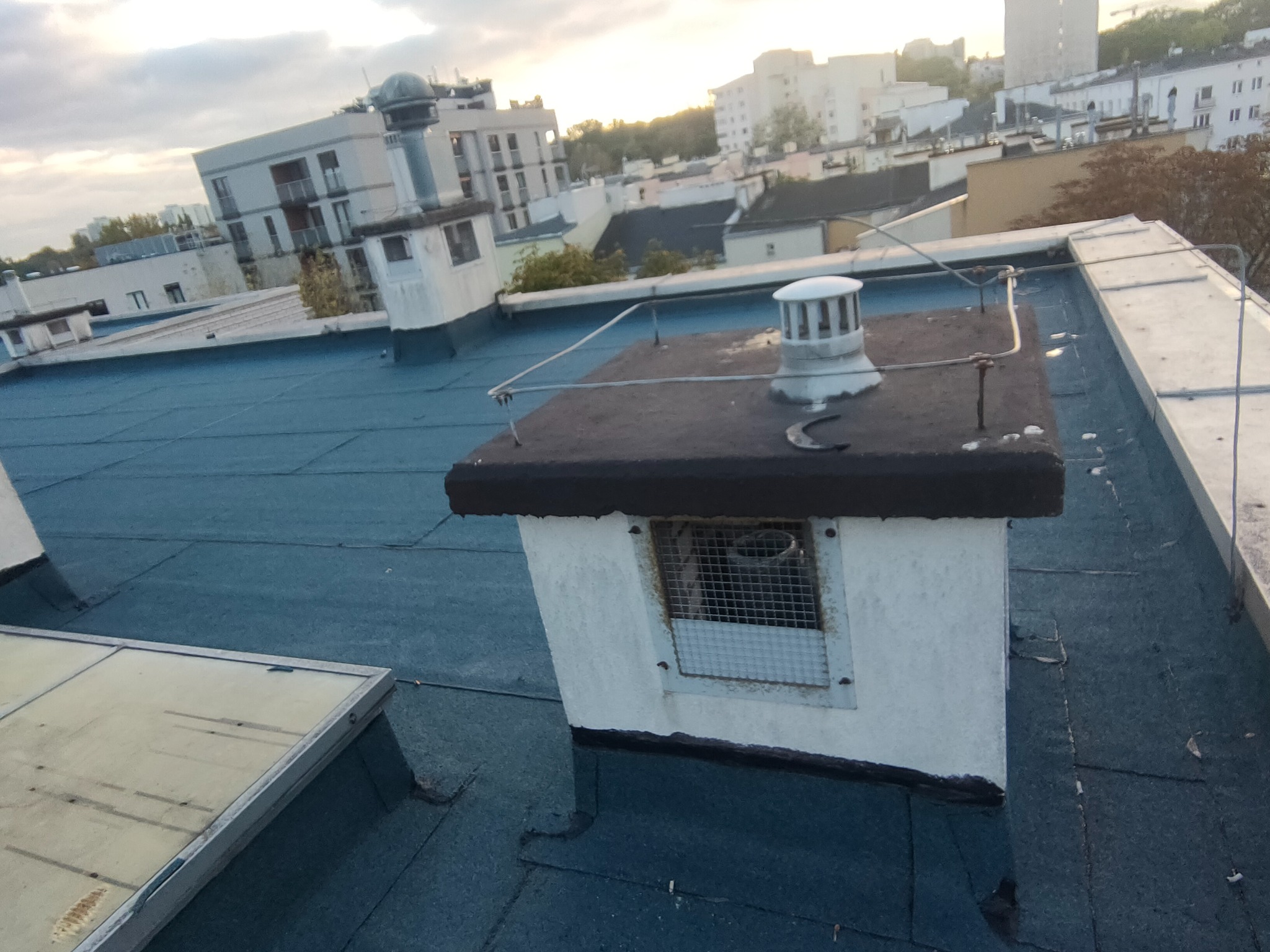 Niebieski dach pokryty papą z kominami wentylacyjnymi i wyłazem dachowym na tle zabudowy miejskiej w pochmurny dzień.