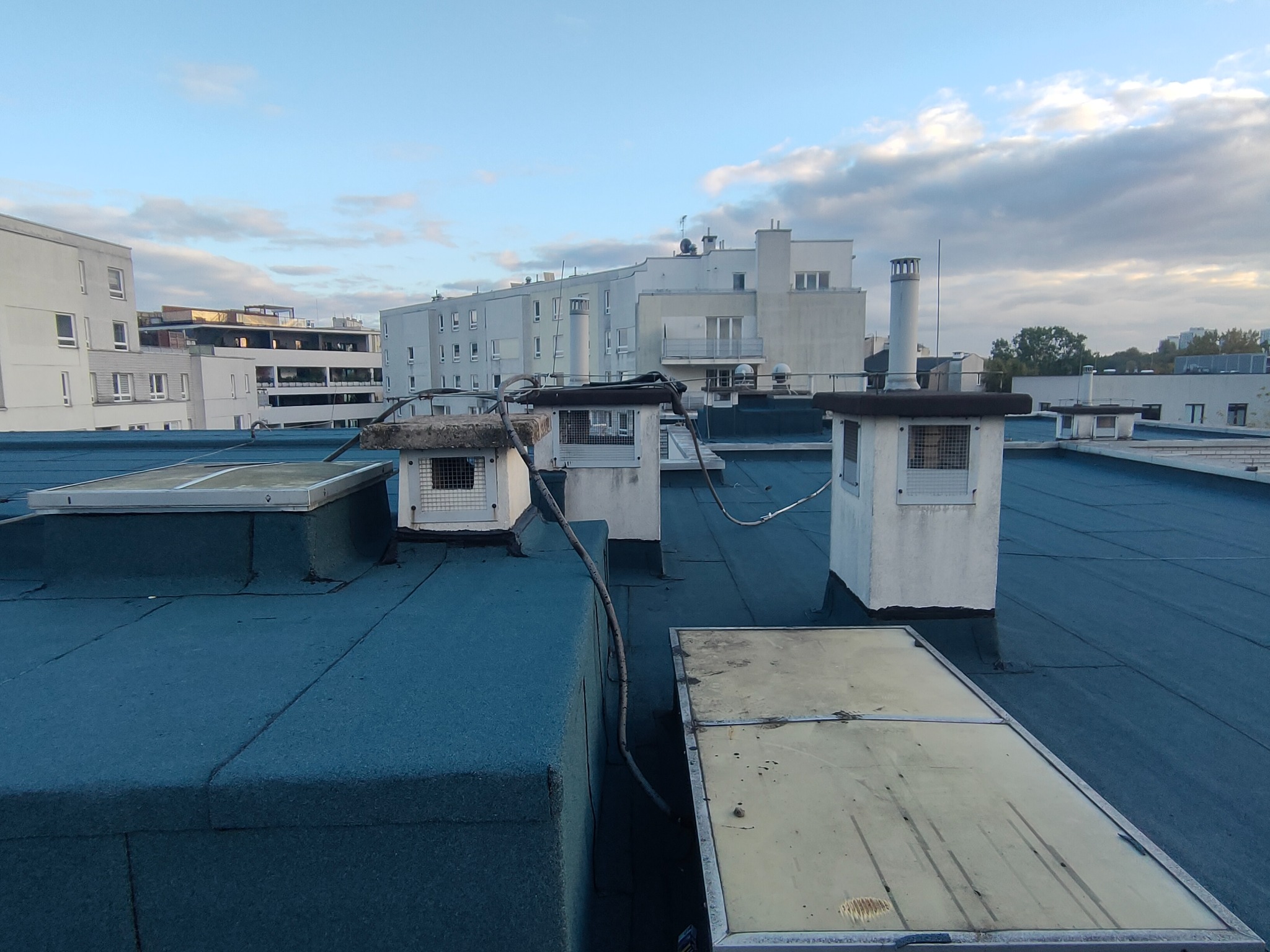 Widok z góry na płaski dach pokryty ciemnoniebieską papą termozgrzewalną z kilkoma wywietrznikami i świetlikami, na tle budynki mieszkalne i pochmurne niebo o poranku.