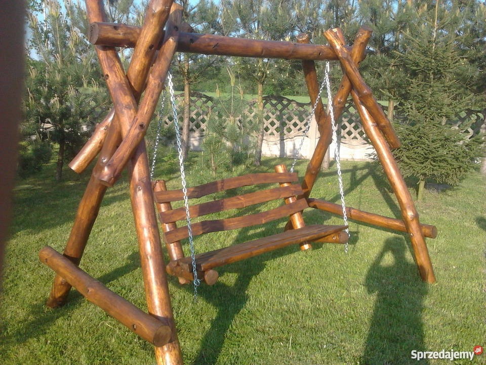 Huśtawka ogrodowa wykonana z okrąglaków drewnianych, zawieszona na metalowych łańcuchach, z widocznym tłem zielonego trawnika i drzew.