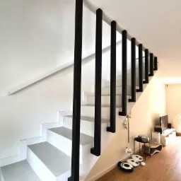 Nowoczesne, minimalistyczne schody z białymi stopniami i czarnymi, kwadratowymi słupkami balustrady, widoczne z dołu do góry, w tle jasne wnętrze domu.