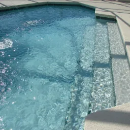 Krystalicznie czysta woda w basenie z delikatnym wodospadem i stopniowanymi schodami wejściowymi, odbijająca promienie słoneczne.