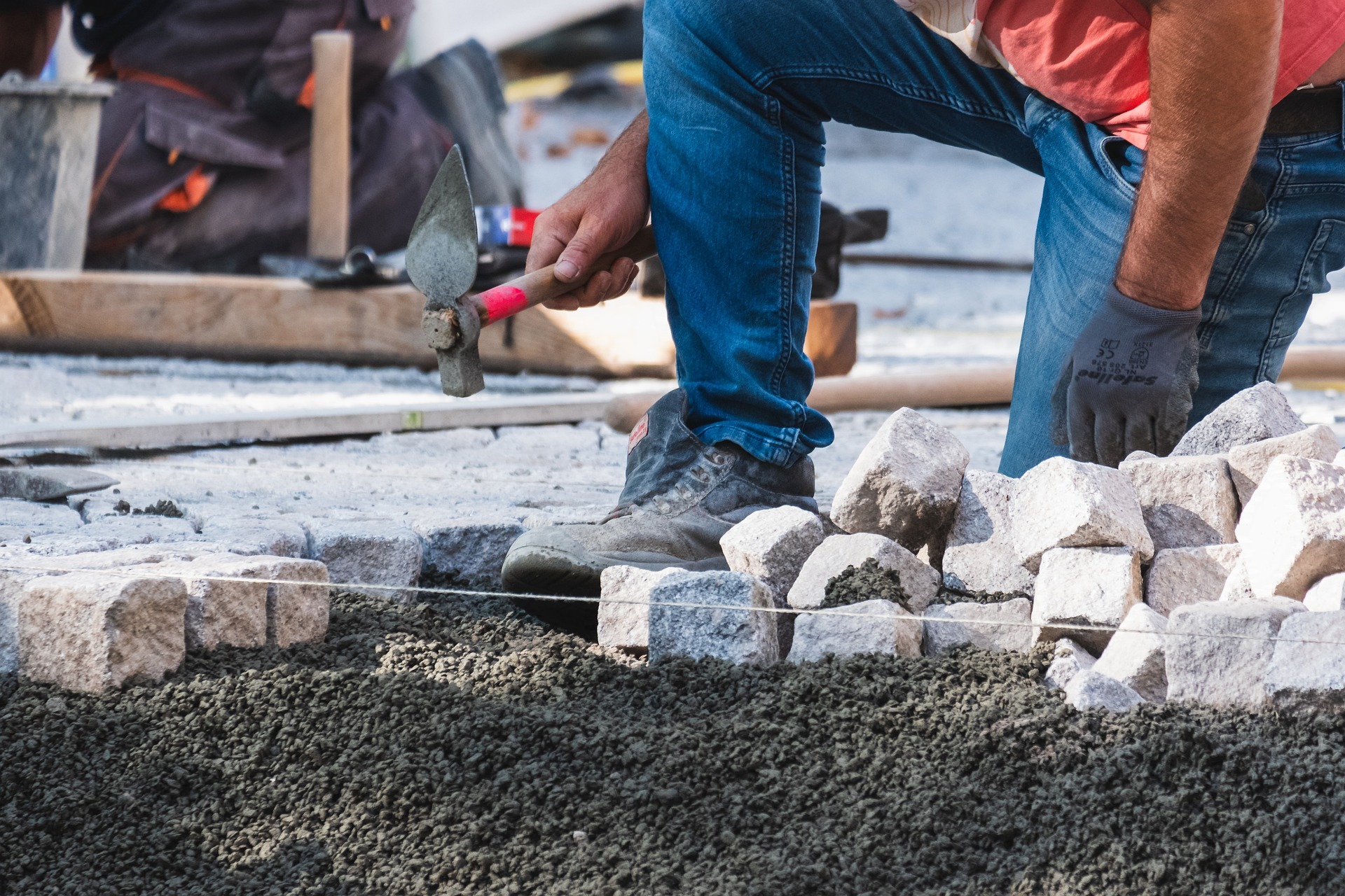 Pracownik w jeansach i rękawicach roboczych dobija młotkiem kamienną kostkę brukową na podsypce, widoczny sznurek wyznaczający linię układania.