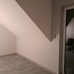 Pokój na poddaszu po remoncie z szarymi panelami podłogowymi, świeżo pomalowanymi ścianami w odcieniach szarości i białymi listwami przypodłogowymi oraz oknem balkonowym.