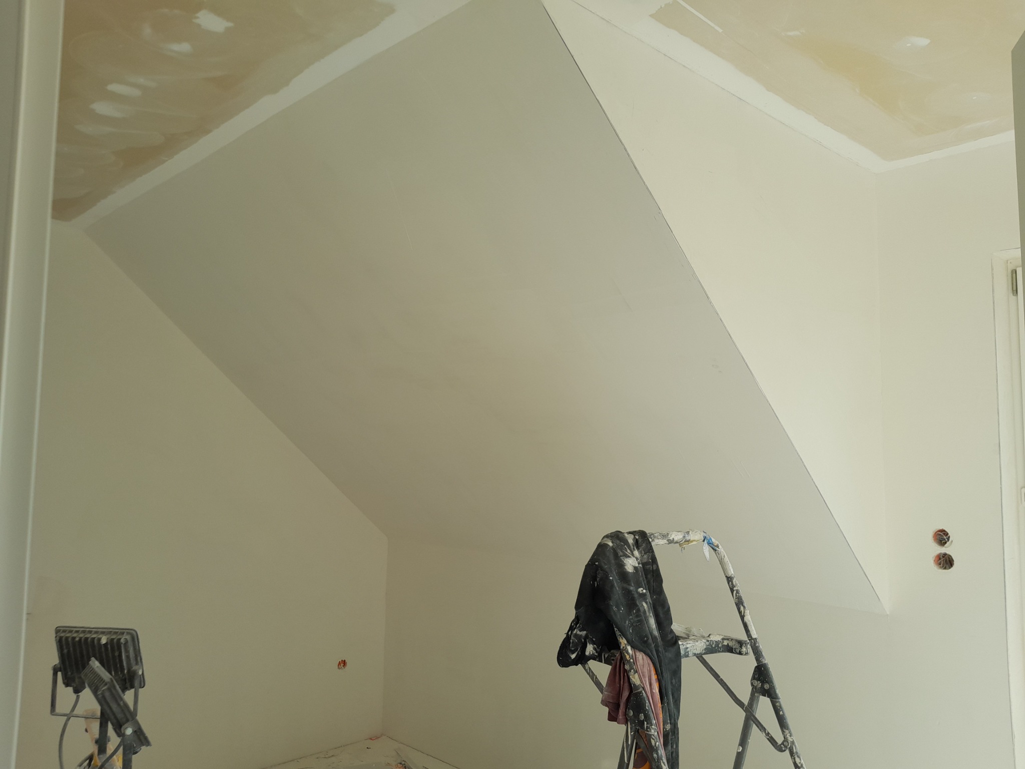 Poddasze w trakcie prac wykończeniowych, widoczna drabina malarska z ubraniami roboczymi, lampa halogenowa i wstępnie wyszpachlowany sufit ze skosem.