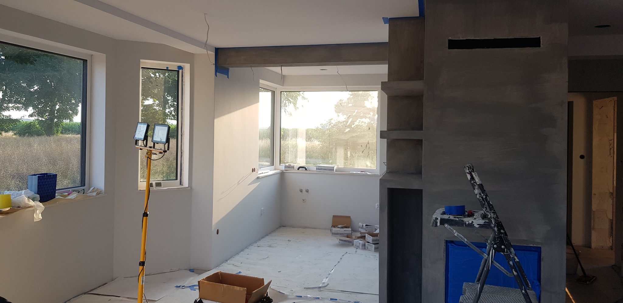 Wnętrze w trakcie prac przygotowawczych do malowania: zabezpieczone okna taśmą malarską, podłoga przykryta folią, widoczny stojak z lampami halogenowymi i drabina malarska przy ścianie z szarą...