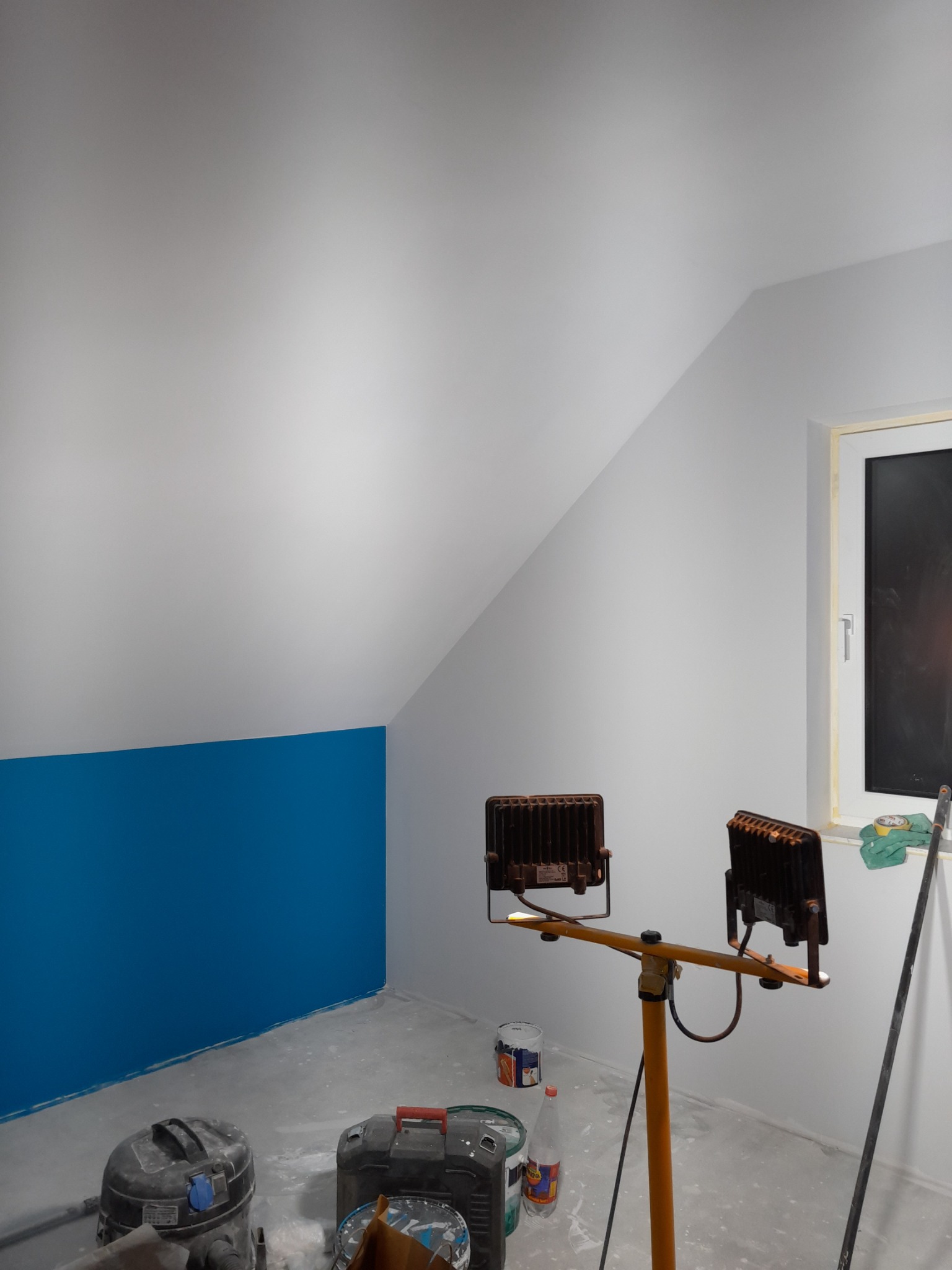 Pokój na poddaszu w trakcie malowania: białe ściany z fragmentem pomalowanym na intensywny niebieski, żółty statyw z lampami halogenowymi oświetlający wnętrze, narzędzia malarskie i akcesoria...