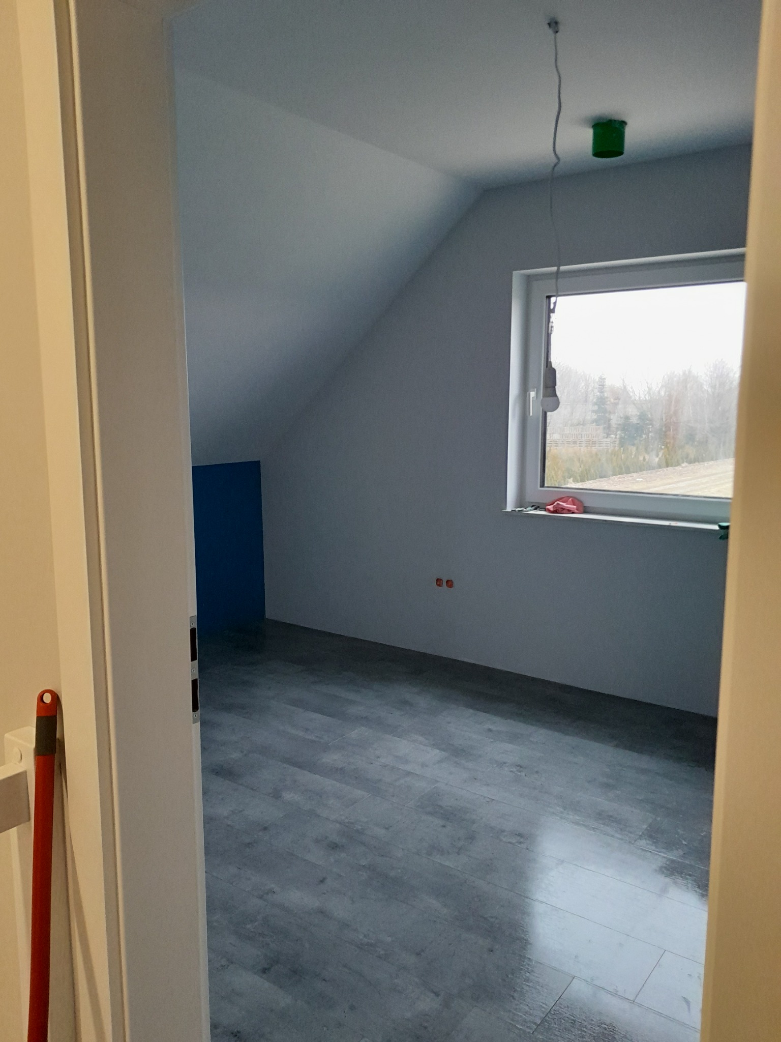 Pokój na poddaszu w trakcie prac wykończeniowych, widoczne szare panele podłogowe, ściany przygotowane do malowania, okno z widokiem na zewnątrz, brak lampy, widoczna instalacja elektryczna.