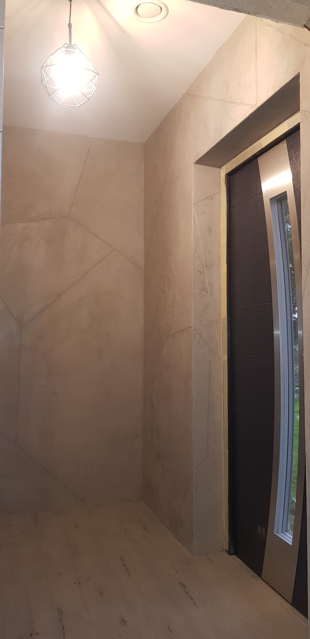 Korytarz z dekoracyjną ścianą w odcieniach beżu, z geometrycznym wzorem przypominającym pęknięcia, oświetlony nowoczesną lampą wiszącą i wbudowanym światłem, z widocznymi fragmentami drzwi...