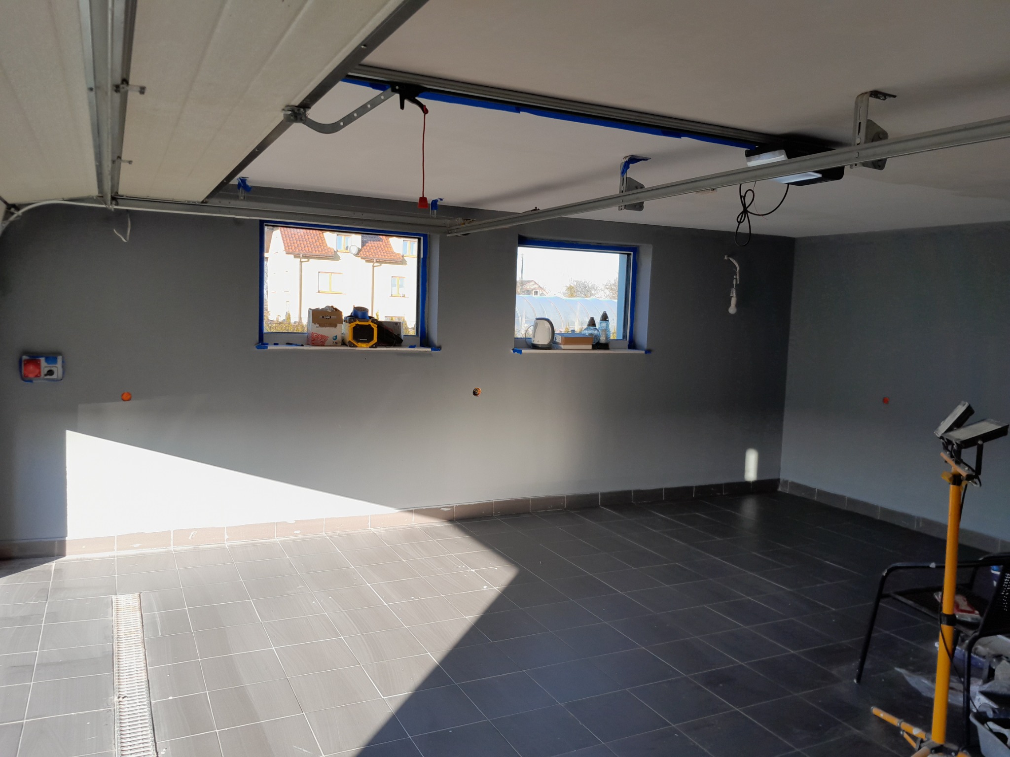Wnętrze garażu w Radymnie po malowaniu ścian na szaro, widoczne okna zabezpieczone taśmą malarską, szara terakota na podłodze, lampa warsztatowa i elementy konstrukcji bramy garażowej pod sufitem.