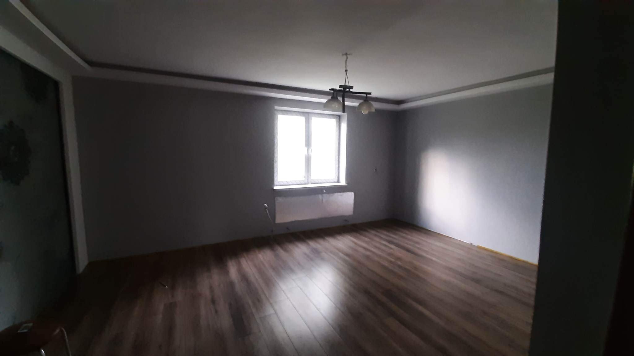 Wnętrze pokoju po remoncie, ściany pomalowane na szaro, podłoga wyłożona panelami, widoczny grzejnik pod oknem i lampa sufitowa z trzema kloszami.