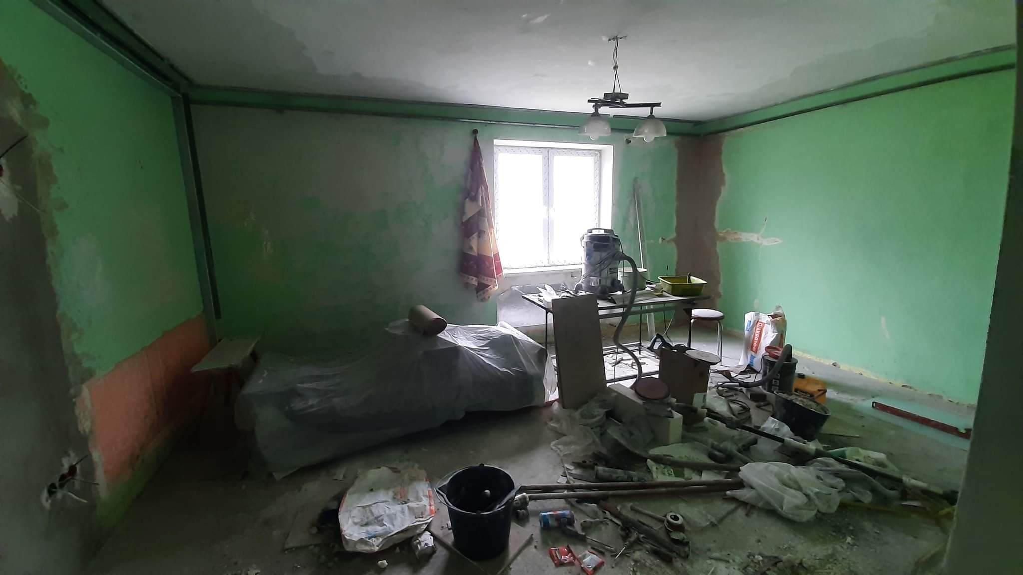 Pokój w trakcie przygotowania do prac wykończeniowych: zielone ściany z fragmentami świeżo nałożonej gładzi, metalowe profile pod sufitem, narzędzia i materiały budowlane na podłodze, okno...