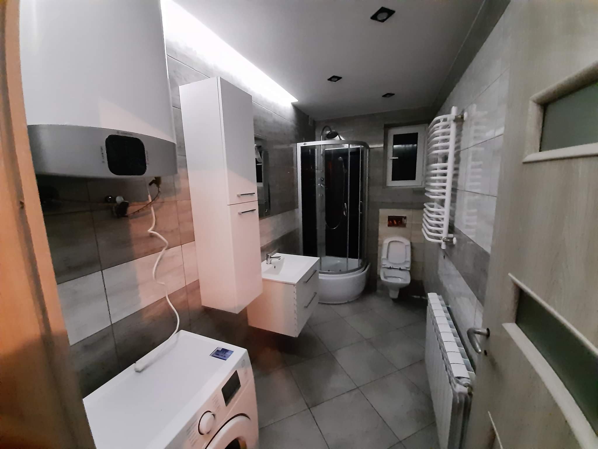 Wykończona łazienka z pralką, kabiną prysznicową, umywalką z szafką i wysoką szafką, grzejnikiem drabinkowym i zwykłym, oświetleniem sufitowym LED i płytkami w odcieniach szarości.