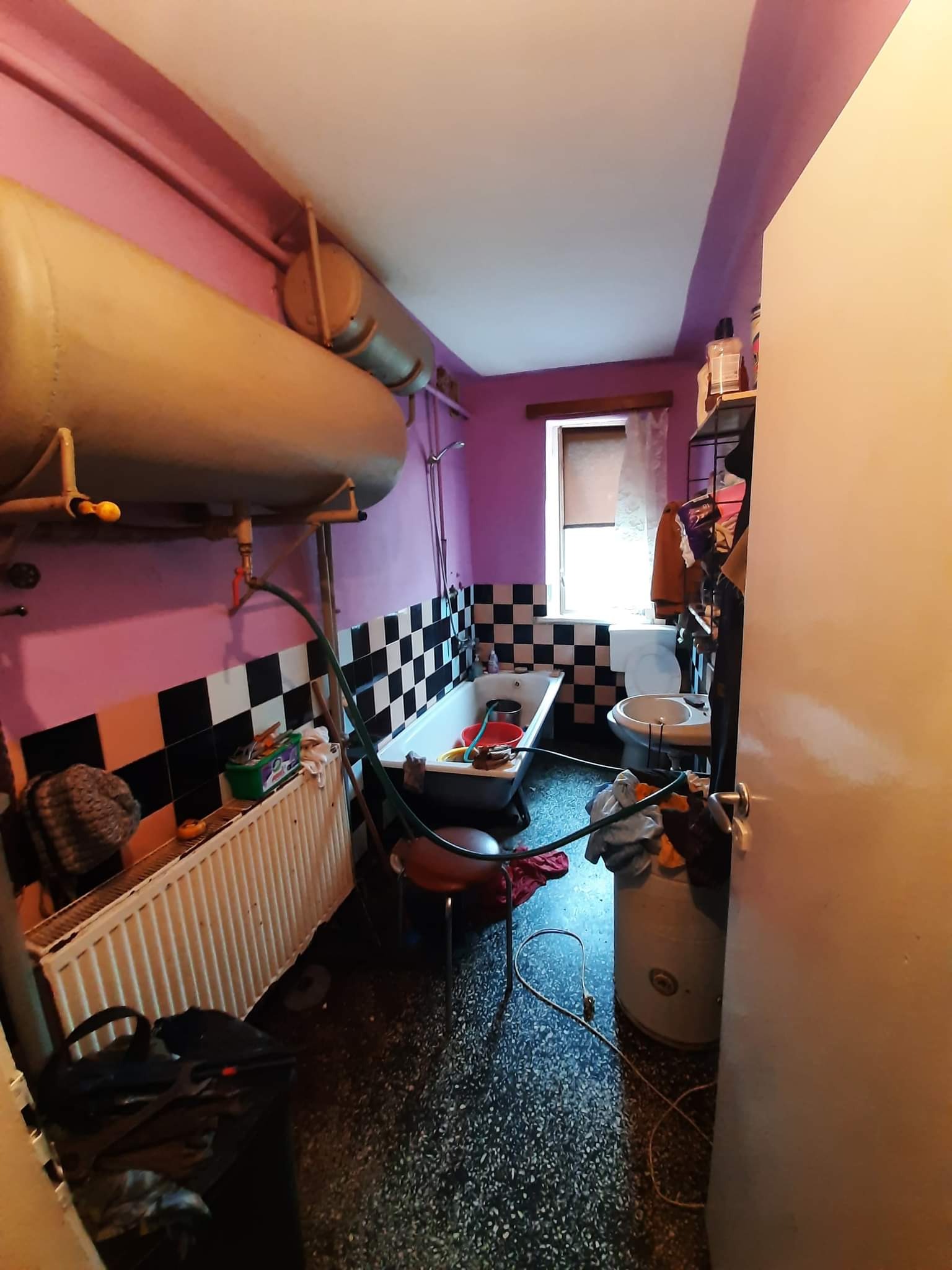 Łazienka przed remontem: różowe ściany, szachownica z płytek, duży bojler na ścianie, bałagan, widoczne ślady użytkowania.