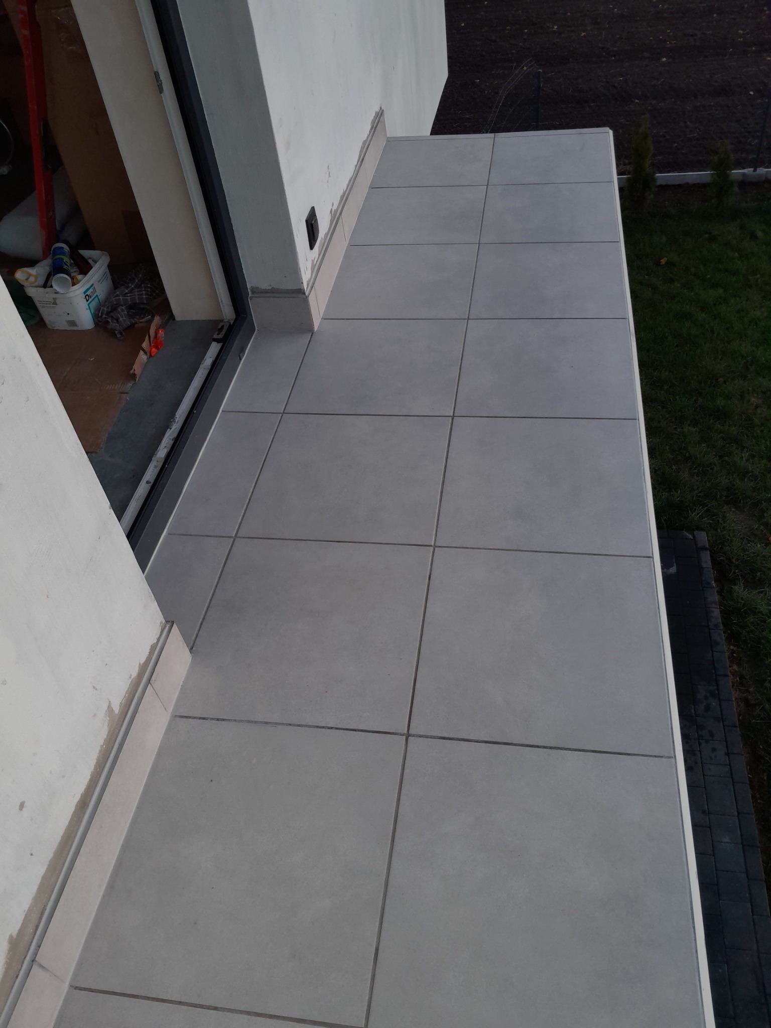Wykończony balkon z szarymi, kwadratowymi płytkami, widok z góry, z widocznymi elementami niezakończonych prac w rogu przy drzwiach balkonowych.