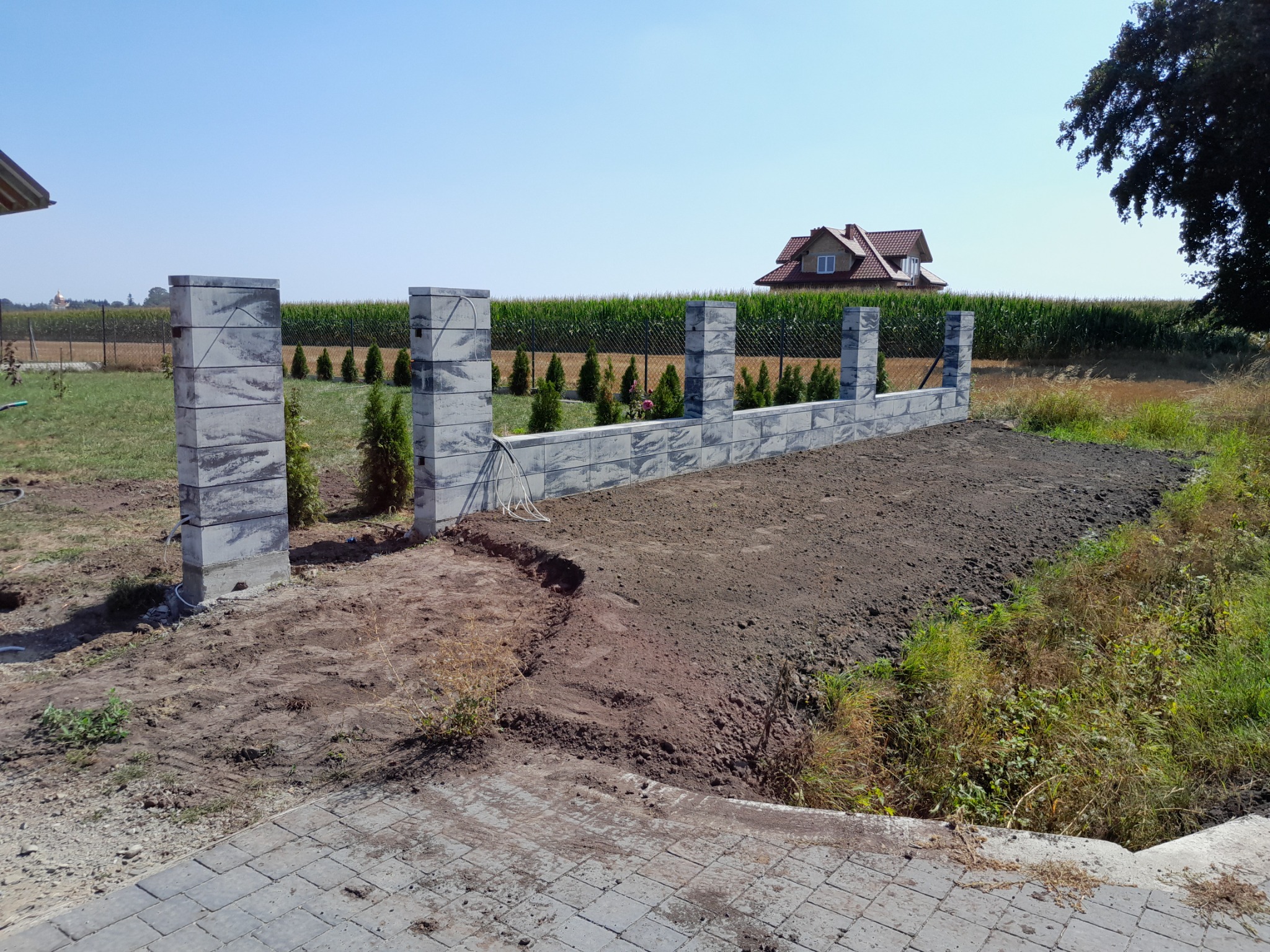 Wznoszenie ogrodzenia z betonowych bloczków w ogrodzie, widoczne słupki i fragment muru, przygotowany teren pod trawnik, w tle dom i pole kukurydzy.