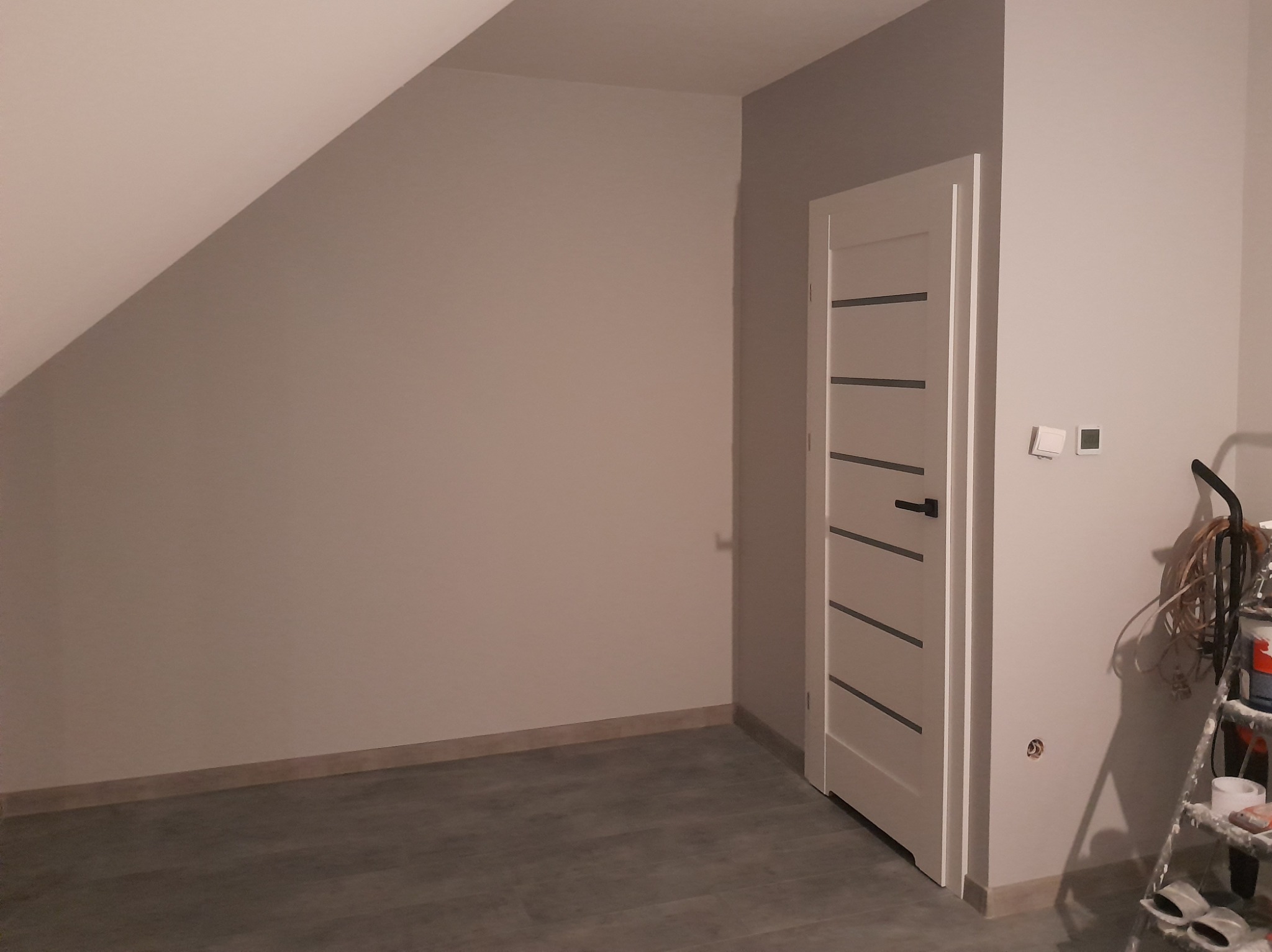 Pokój na poddaszu po odświeżeniu, z pomalowanymi ścianami na jasny odcień szarości, białe drzwi z poziomymi paskami, szare panele podłogowe, widoczne ślady prac malarskich oraz drabina z farbami.