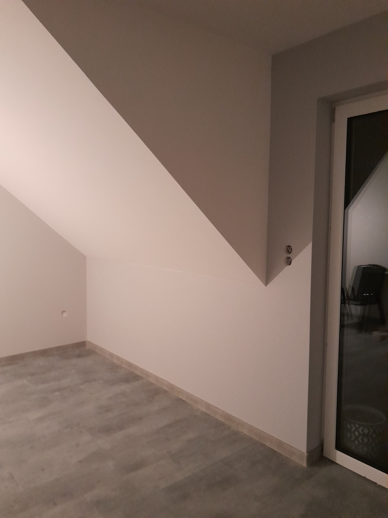 Pokój na poddaszu po remoncie z szarymi panelami podłogowymi, świeżo pomalowanymi ścianami w odcieniach szarości i białymi listwami przypodłogowymi oraz oknem balkonowym.