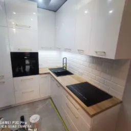 Nowoczesna kuchnia z białymi, lakierowanymi frontami szafek, złotymi uchwytami, drewnianym blatem i białymi płytkami ściennymi typu metro, z widoczną płytą indukcyjną i zlewem, częściowo...