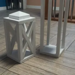 Dwie białe drewniane latarnie dekoracyjne, jedna z krzyżowym wzorem, druga z uchwytem ze sznura, stojące na podłodze imitującej drewno w pomieszczeniu z rośliną doniczkową w tle.