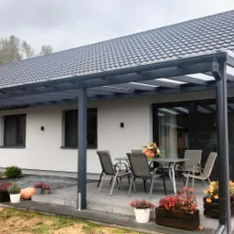Nowoczesna pergola z metalową konstrukcją przylegająca do domu, z szarymi meblami ogrodowymi i dekoracjami z dyni i kwiatów w donicach.