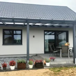 Nowoczesny taras z zadaszeniem z szarej konstrukcji, połączony z domem, z meblami ogrodowymi i kwiatami w donicach, na tle zadbanego ogrodu.