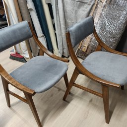 Tapicer - Dwa krzesła z drewnianą ramą i szarą tapicerką stoją obok siebie, widoczne materiały tapicerskie w rolkach w tle.