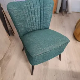 Turkusowy fotel z tapicerowanym oparciem w stylu retro, z ciemnymi, drewnianymi nóżkami, na jasnej podłodze.