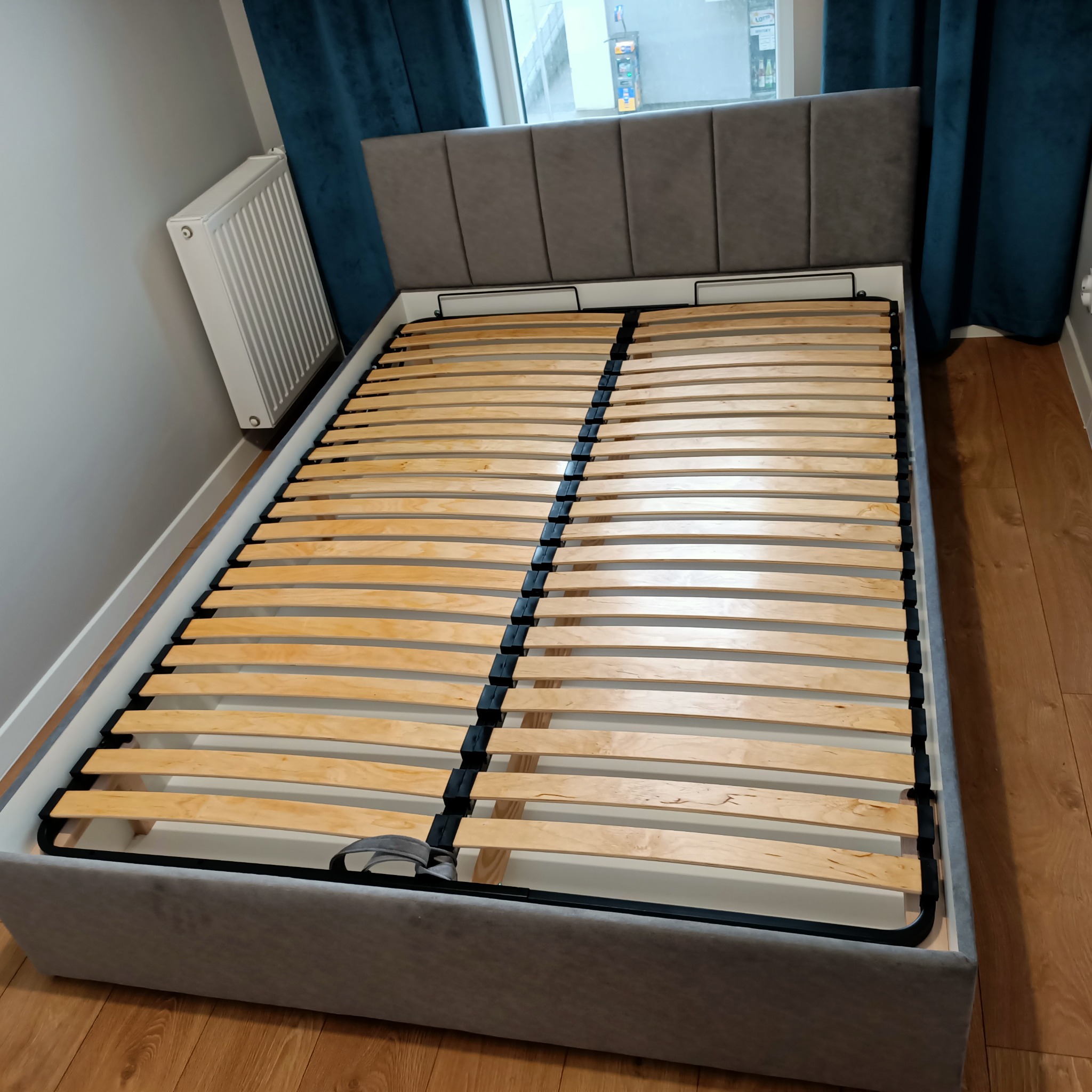 Szare tapicerowane łóżko z drewnianym stelażem listwowym, widok z góry na ramę i zagłówek z podziałem na panele.