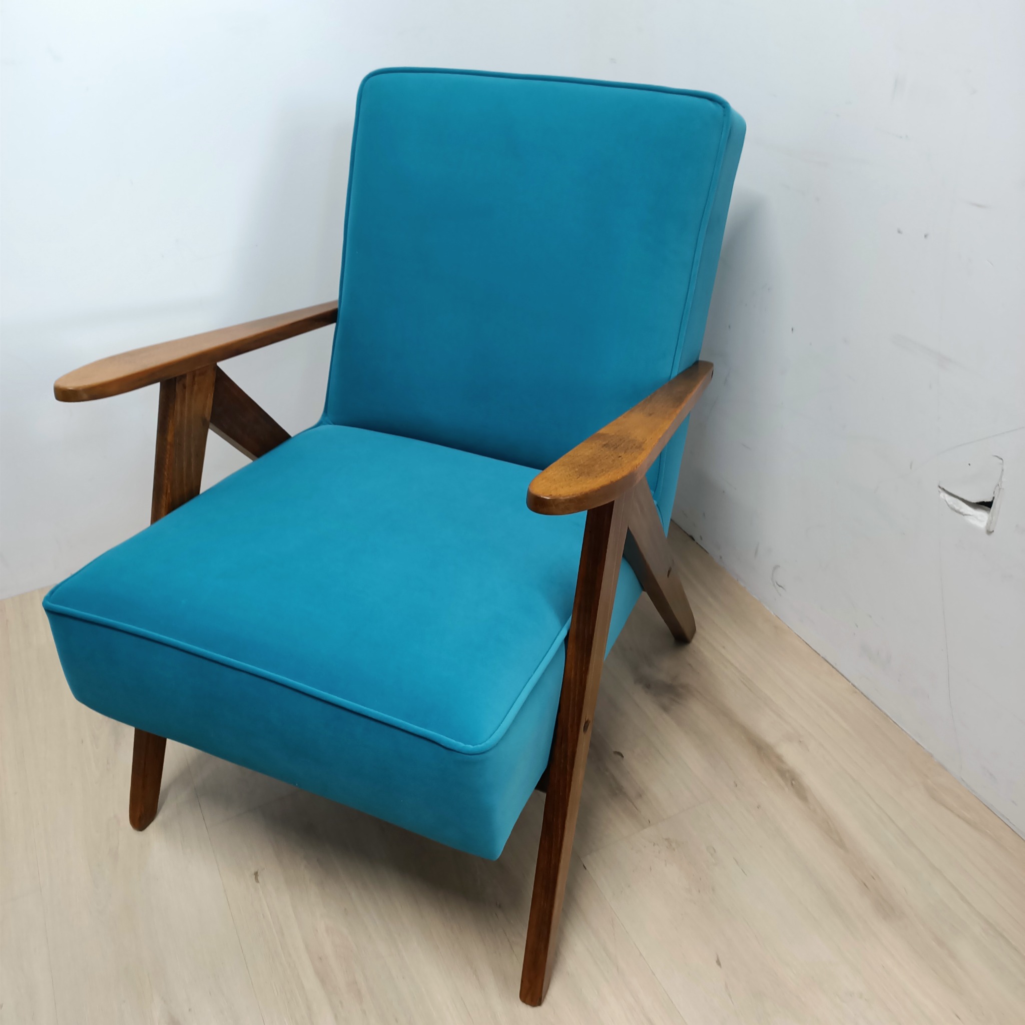 Turkusowy fotel z drewnianymi podłokietnikami w stylu vintage, ustawiony na jasnej podłodze i na tle białej ściany z widocznym ubytkiem.