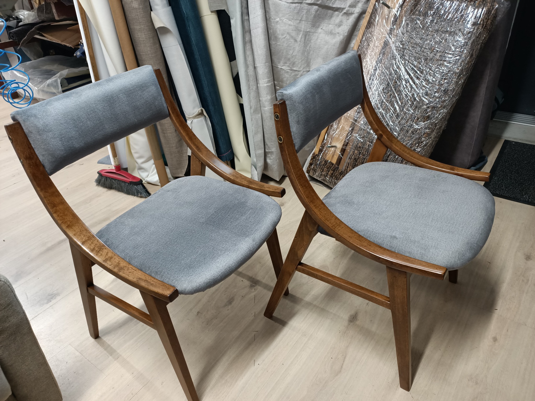 Dwa krzesła z drewnianą ramą i szarą tapicerką stoją obok siebie, widoczne materiały tapicerskie w rolkach w tle.