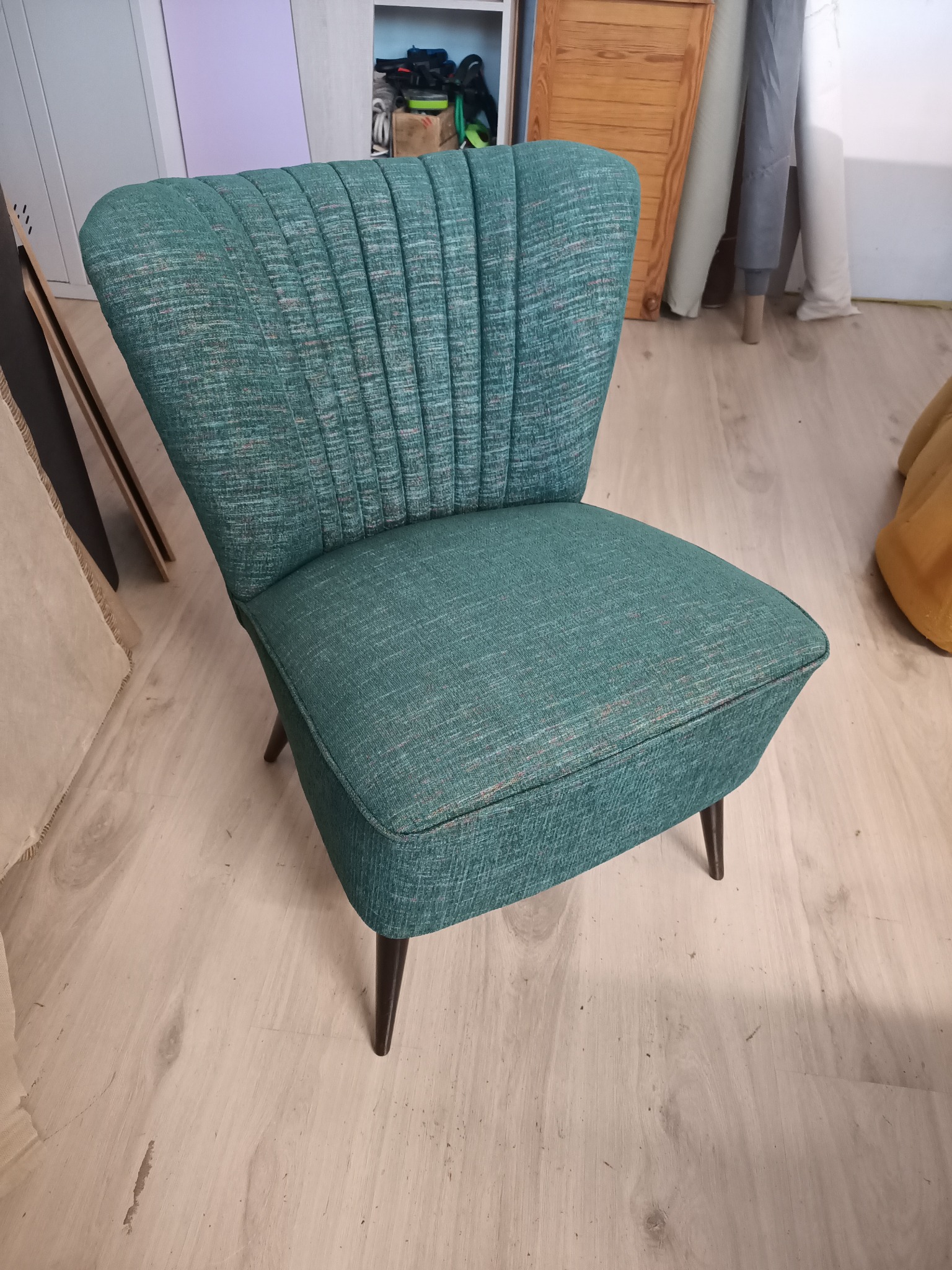 Turkusowy fotel z tapicerowanym oparciem w stylu retro, z ciemnymi, drewnianymi nóżkami, na jasnej podłodze.