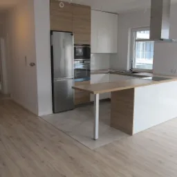 Nowoczesna kuchnia w jasnych barwach z wyspą, drewnianymi akcentami i sprzętem AGD ze stali nierdzewnej, widok na otwartą przestrzeń salonu z parkietem.