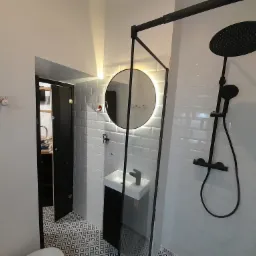 Nowoczesna łazienka z czarną armaturą: deszczownica, umywalka z szafką, okrągłe lustro z podświetleniem LED i wzorzysta podłoga.