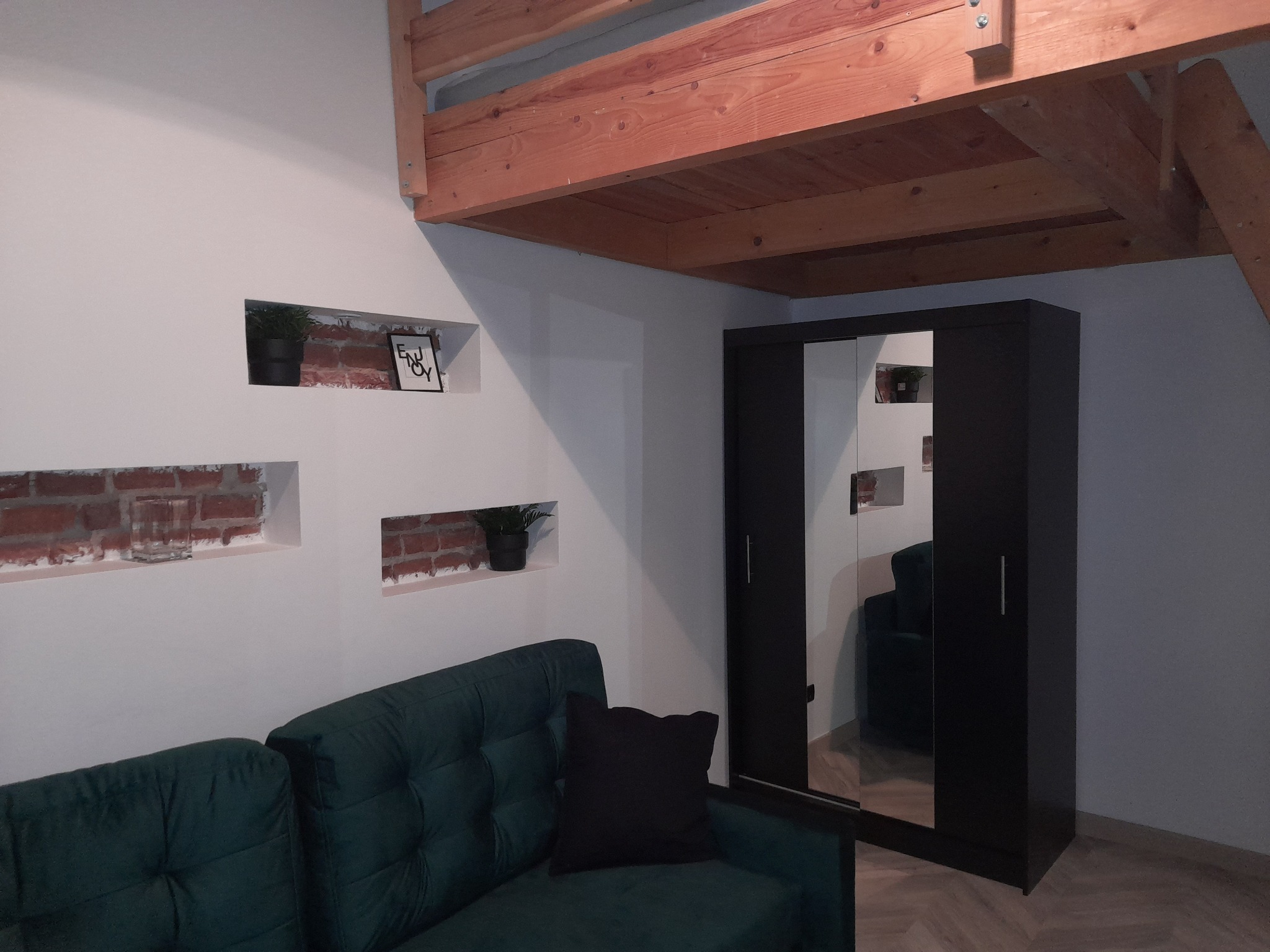 Pokój z antresolą, zieloną sofą i czarną szafą z lustrami, ściana z wnękami wykończonymi czerwoną cegłą, w których umieszczono rośliny i ramkę z napisem 'enjoy'.
