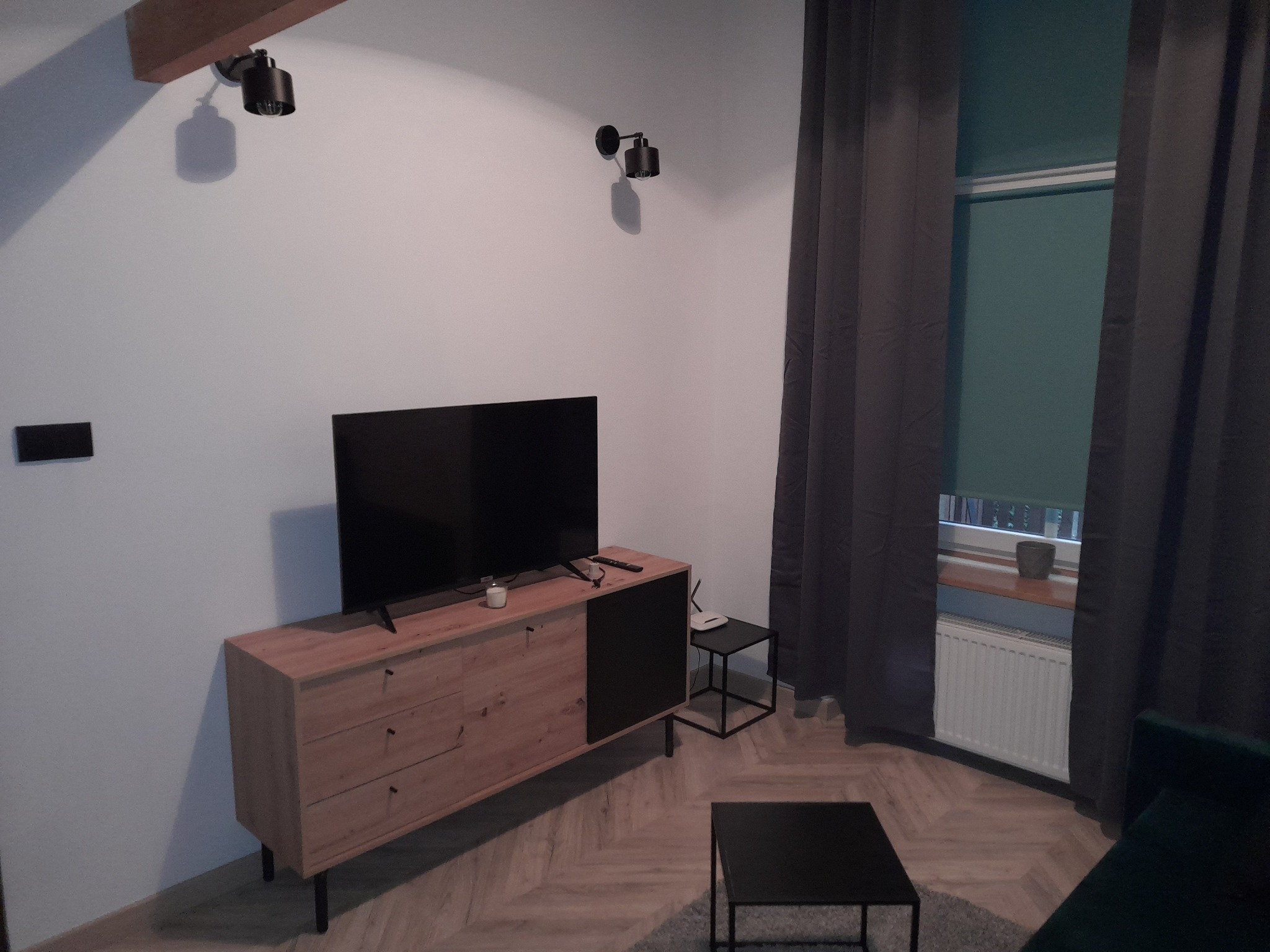 Nowoczesny salon z drewnianą komodą pod telewizorem, minimalistycznymi stolikami kawowymi i szarymi zasłonami, oświetlony czarnymi lampami punktowymi.