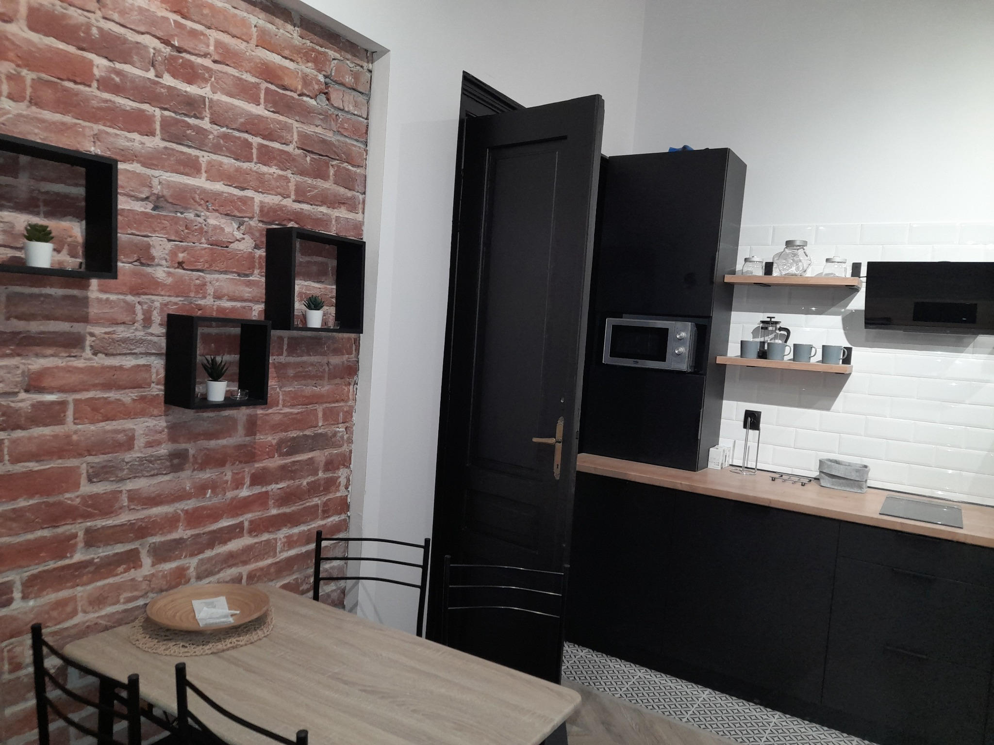 Nowoczesna kuchnia z ceglaną ścianą, czarnymi meblami i drewnianym stołem z dekoracjami, widoczna mikrofalówka i białe płytki na ścianie.