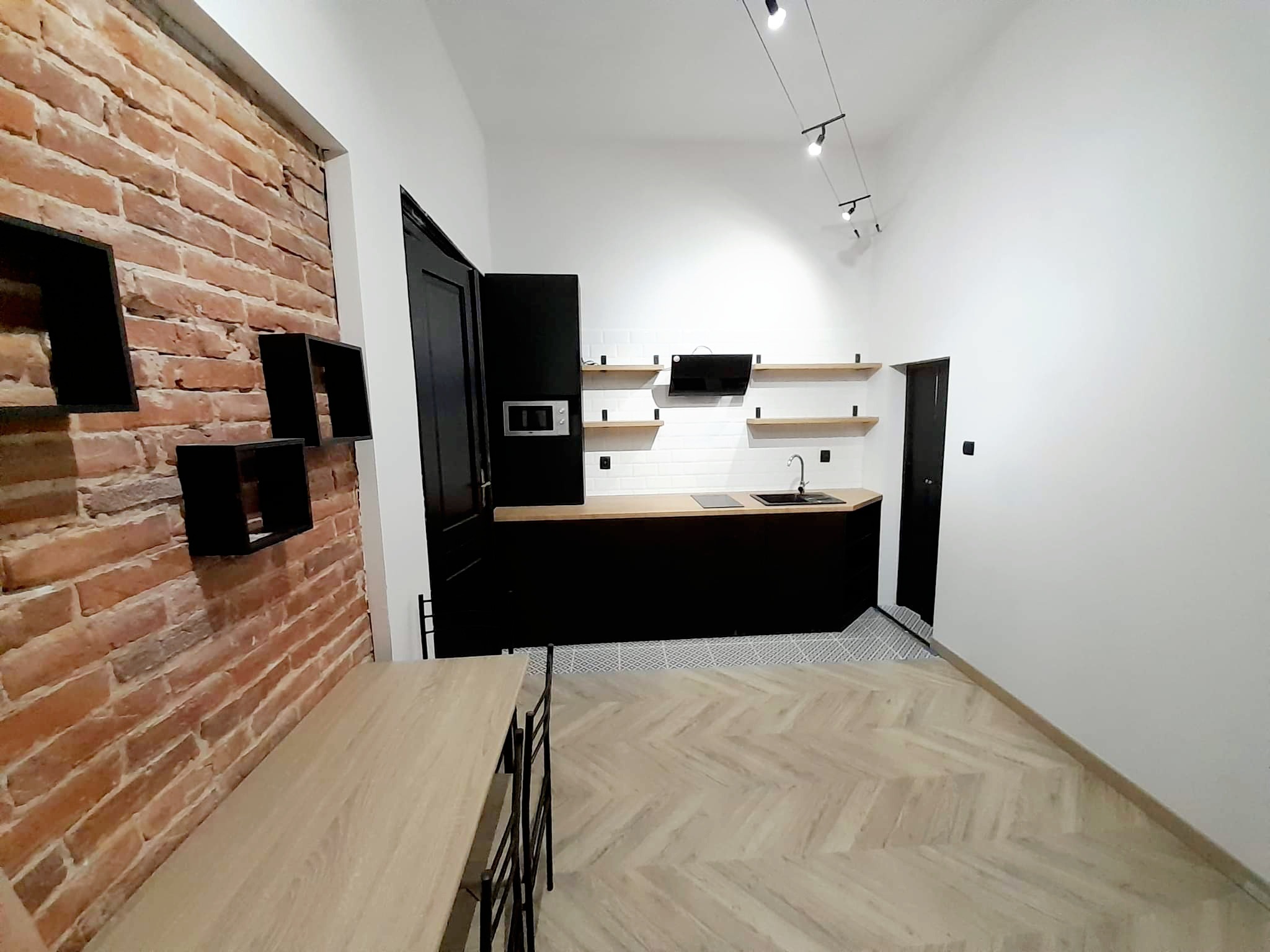 Wnętrze kuchni z ceglaną ścianą, czarnymi szafkami i drewnianym blatem, oświetleniem szynowym i podłogą ułożoną w jodełkę.