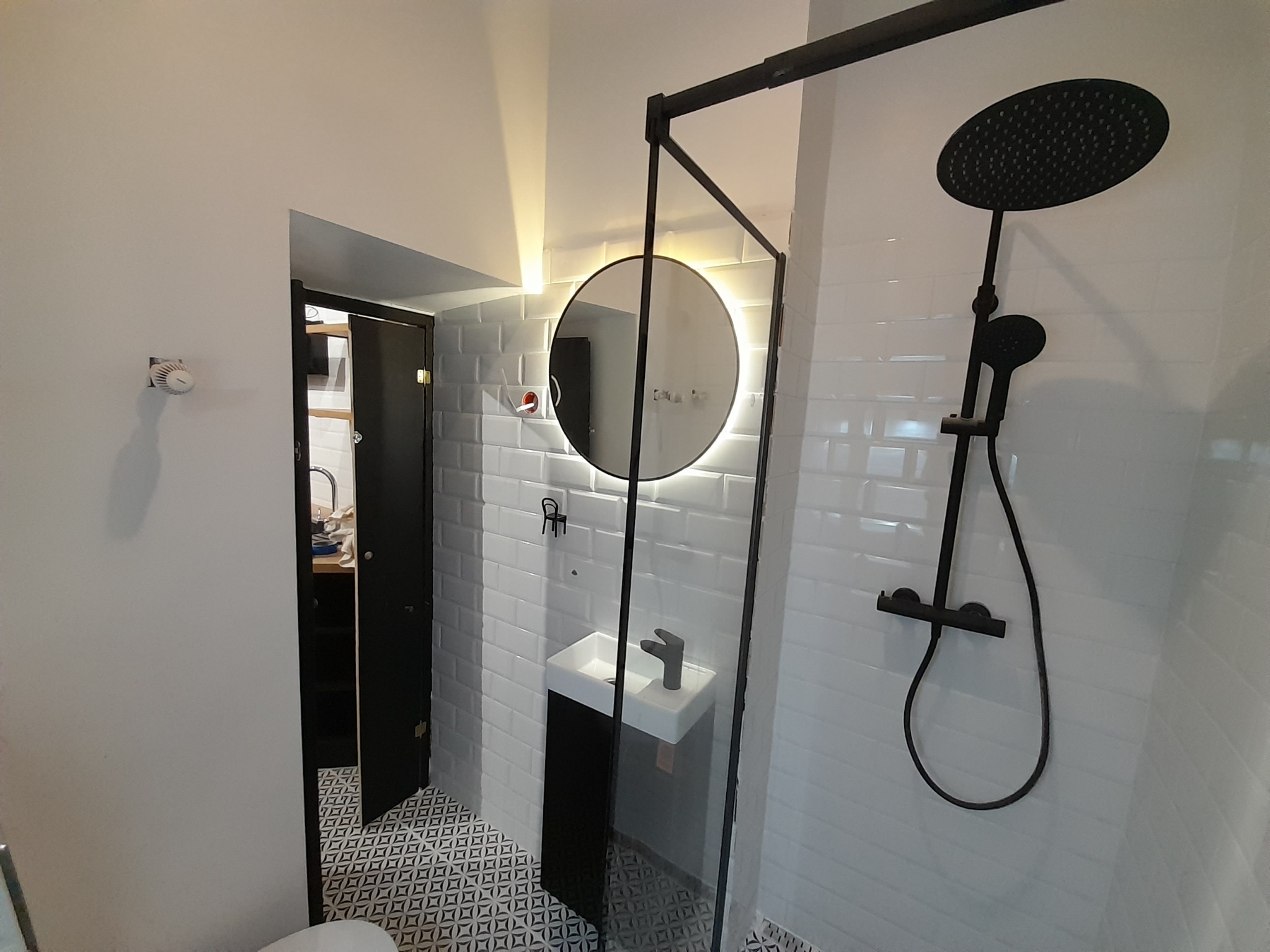 Nowoczesna łazienka z czarną armaturą: deszczownica, umywalka z szafką, okrągłe lustro z podświetleniem LED i wzorzysta podłoga.